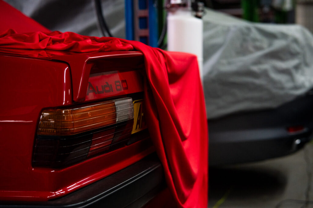 Close-up of a red classic Audi 80 rear light cluster and boot spoiler being uncovered from a silk indoor car cover, representing secure, premium classic car storage.